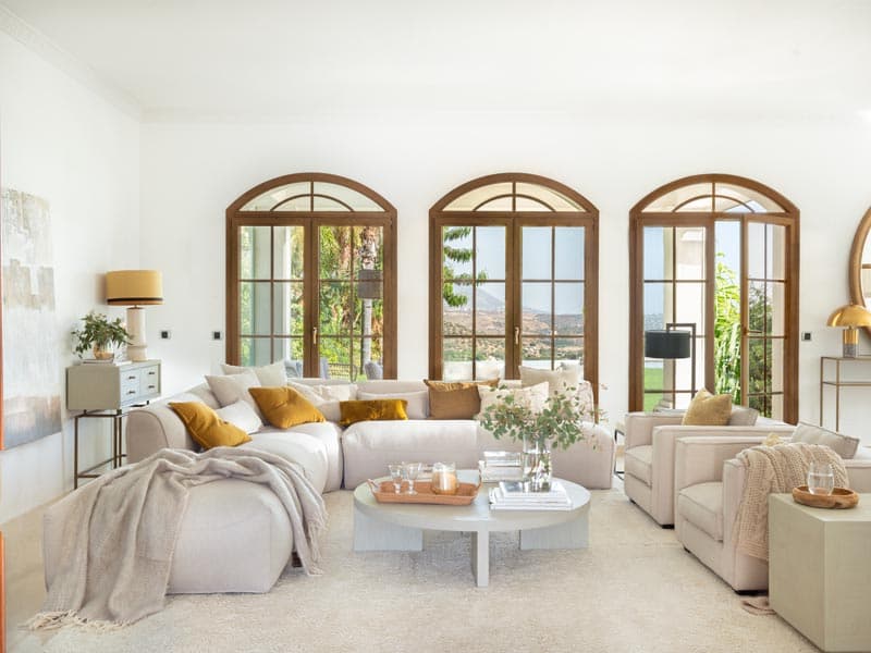 Salón con sofá rinconera de gran formato en beige claro, cojines en mostaza y ocre, mesita de café ovalada blanca y una lámpara de sobremesa dorada, con los característicos ventanales en arco.