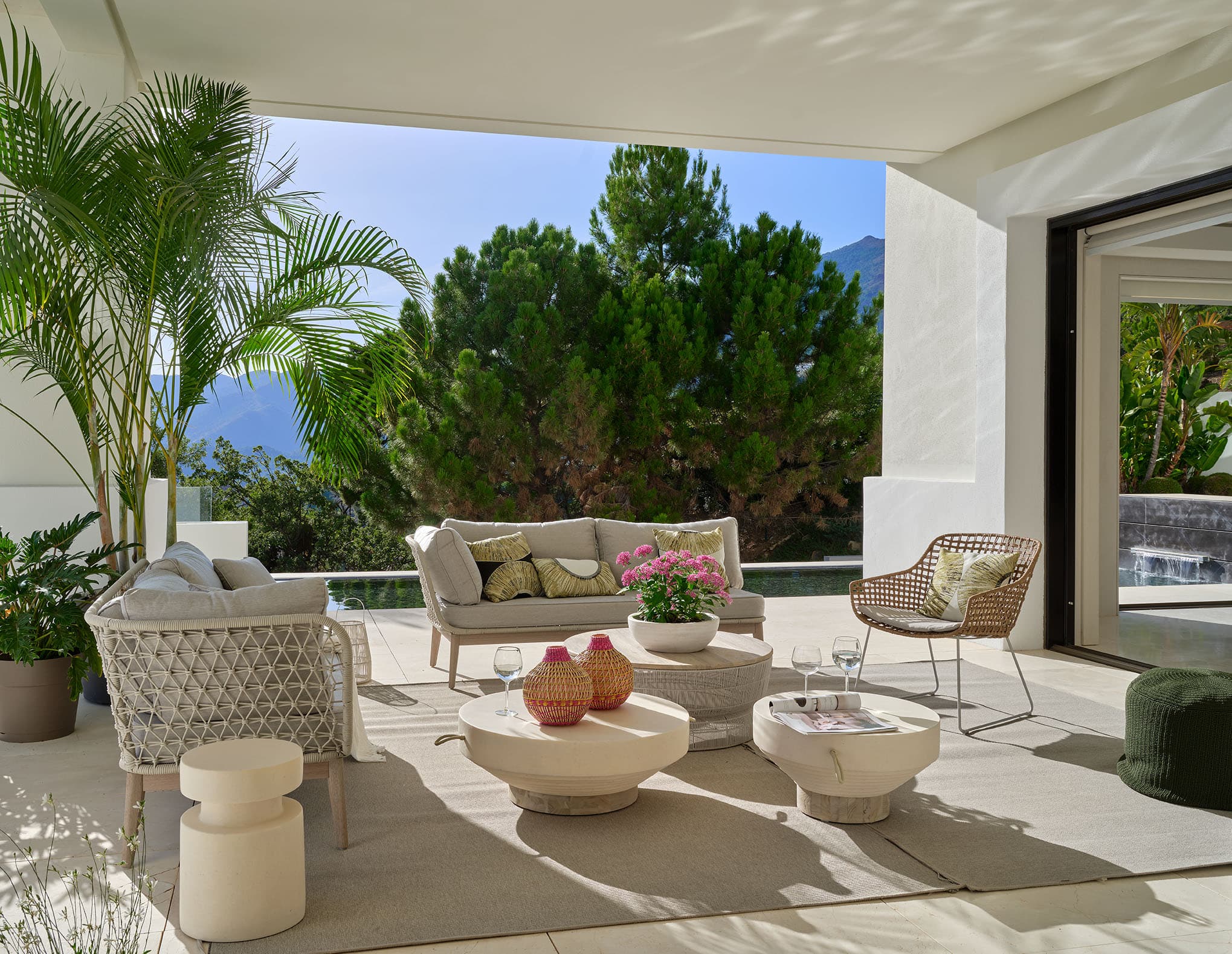 Terraza exterior luminosa de la villa con sillones y sofás de aluminio y cuerda en blanco, mesas de café bajas, decoración en rosa y verde, y vistas a los pinos y la montaña.