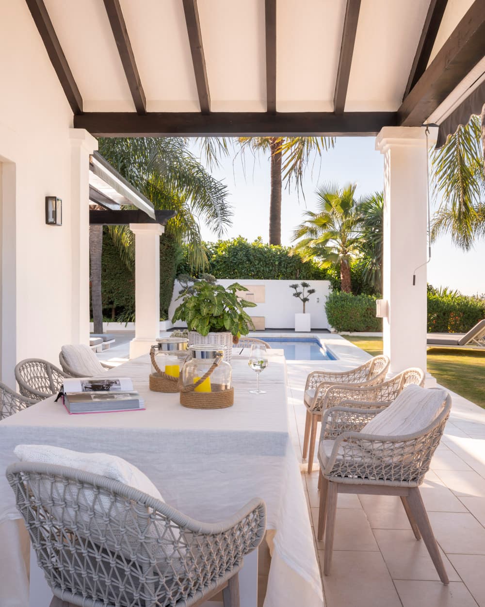 Vista de la terraza cubierta con mesa de lino blanca y sillas de cuerda trenzada gris, con columnas blancas y vigas oscuras enmarcando las palmeras tropicales del jardín y la piscina al fondo. El ambiente destila el refinamiento y la ligereza de la vida al aire libre en el sur de España.