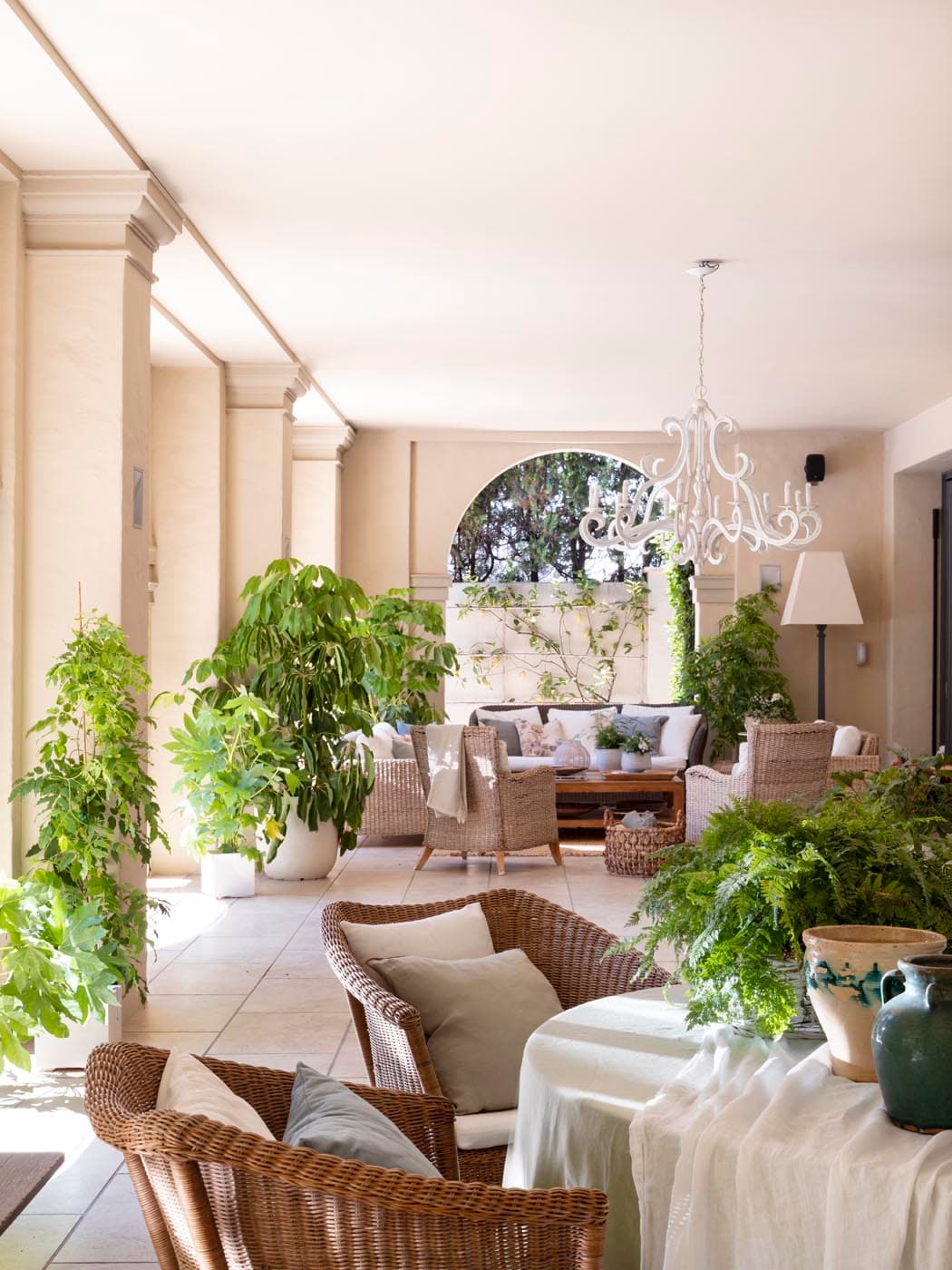 Galería porticada interior de gran amplitud con columnas clásicas, suelo de baldosa grande y techo liso con araña de hierro forjado blanco. La estancia está repleta de plantas tropicales: ficus, filodendros y helechos en grandes macetas. Sillones y butacas de mimbre beige crean zonas de estar informales. Al fondo, un arco da paso al jardín.