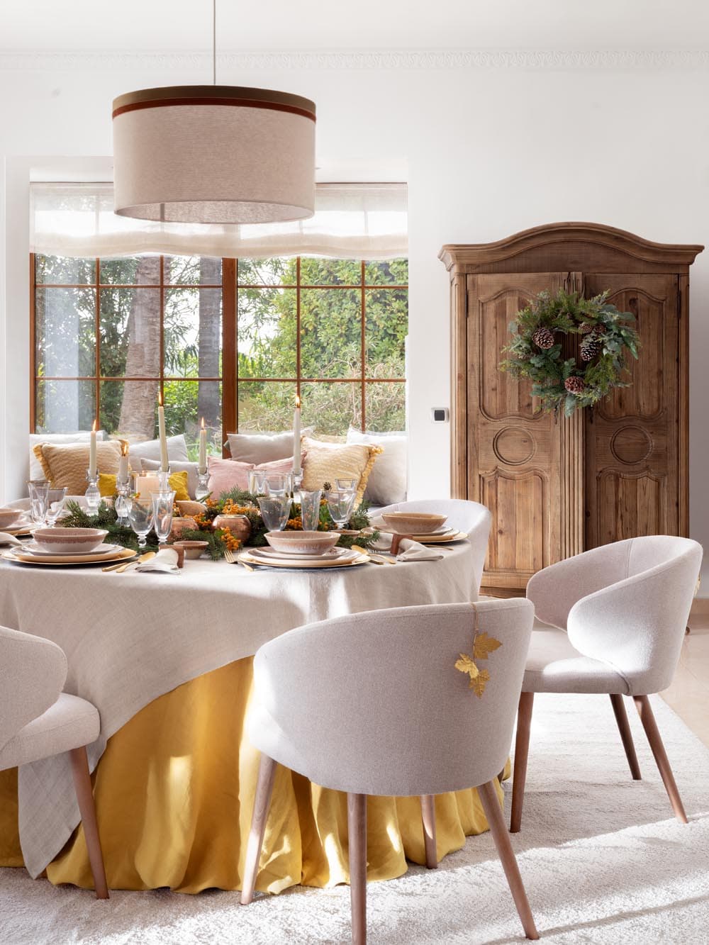 Vista del comedor navideño con gran armario provenzal de madera natural coronado por una corona de pinos y bayas. La mesa redonda aparece con vajilla artesanal, velas y centro de ramas de pino y piñas, junto a un banco corrido tapizado con cojines en tonos cálidos al fondo.