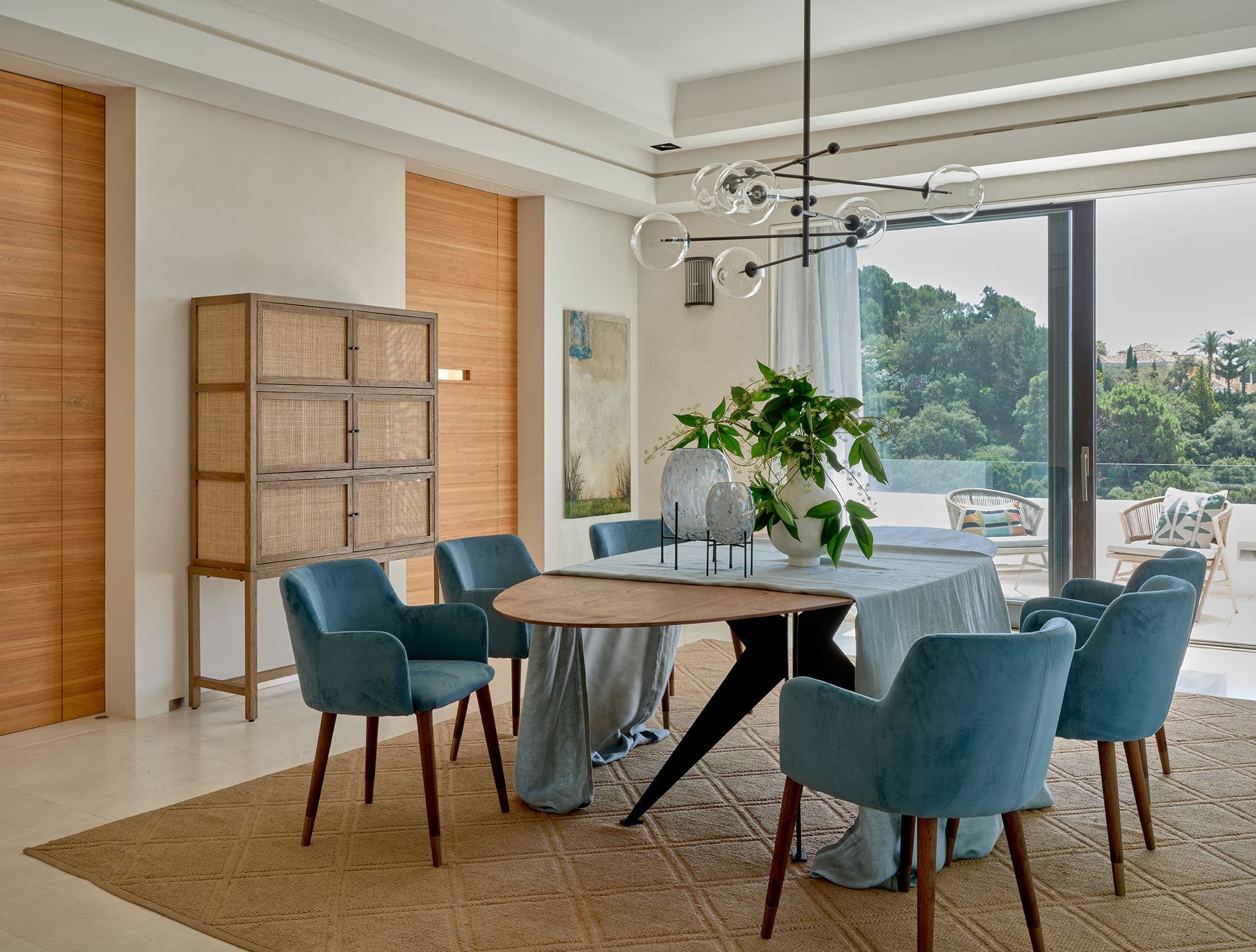 Comedor interior de diseño contemporáneo con mesa de madera orgánica, sillas tapizadas en terciopelo azul-verde, lámpara de burbujas de vidrio y armario de ratán en madera natural.