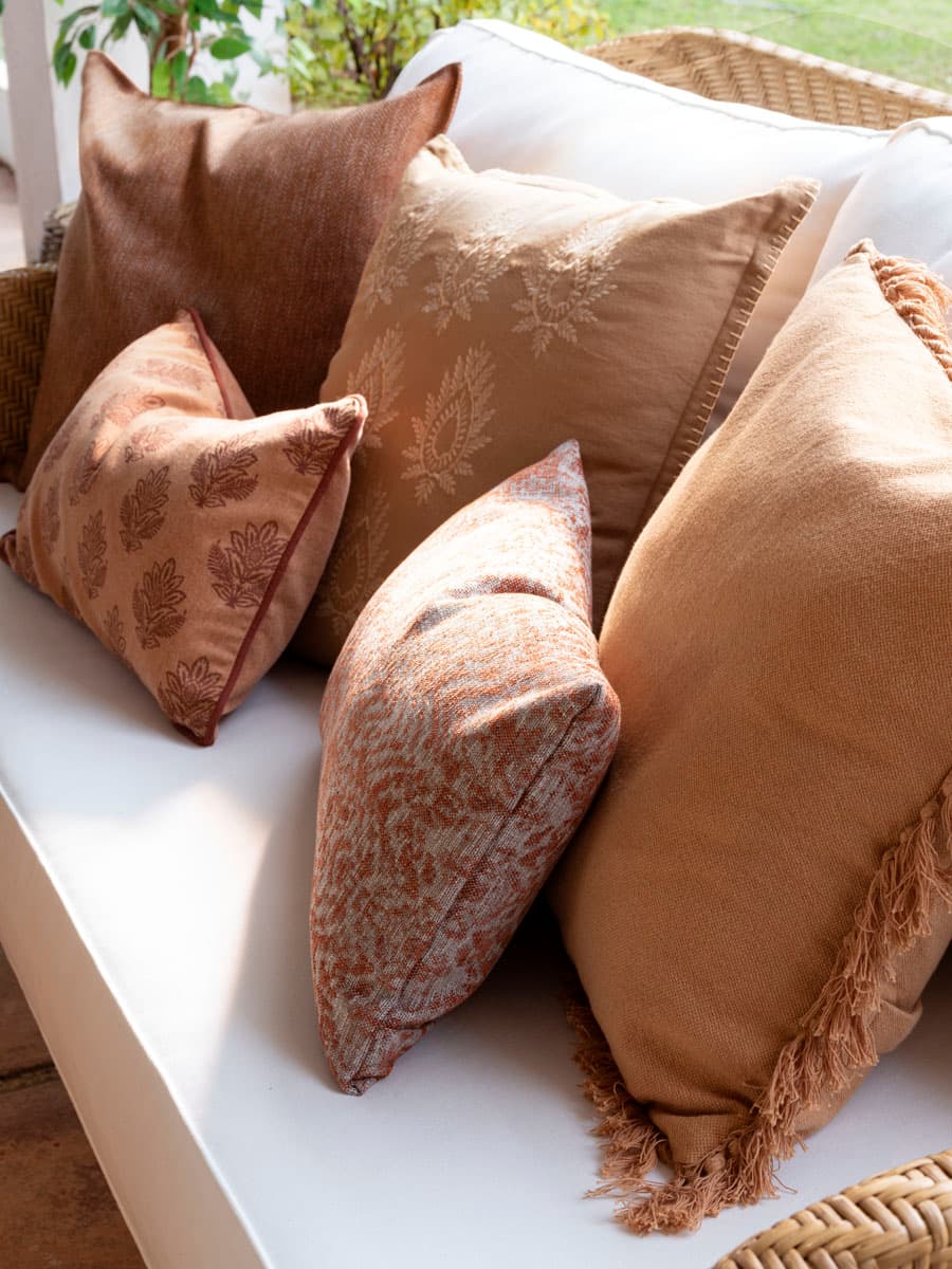 Detalle de los cojines decorativos apilados sobre el sofá de ratán. La selección combina cojines en tonos tierra: marrón chocolate, beige arena, terracota oxidado y ocre, con texturas y estampados variados —tejido plano, motivos florales blockprint, estampado vegetal difuminado y flecos en los bordes—. La paleta cálida y terrosa es coherente con el carácter orgánico y mediterráneo del proyecto.