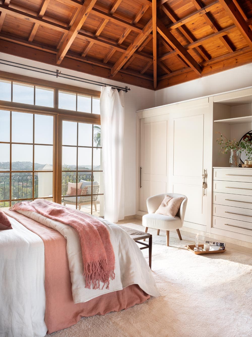 Vista del dormitorio principal enfocada en la zona de vestidor con armarios empotrados lacados en blanco y cómoda integrada, junto a la balconera de madera con vistas a la sierra y terraza. La butaca en bouclé y la manta de cachemira en rosa crean un rincón íntimo y elegante.