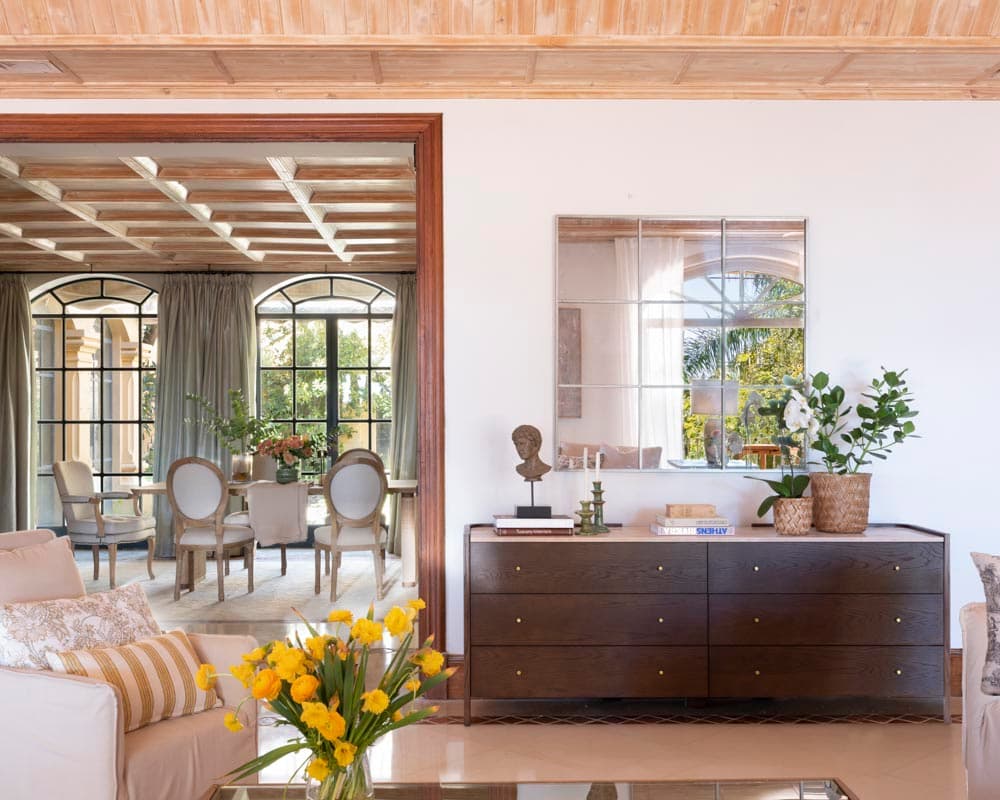 Rincón del salón con una cómoda de madera oscura decorada con busto clásico y orquídea blanca, sobre la que cuelga un espejo de cuarterones metálico. Al fondo, a través del arco de madera oscura, se vislumbra el despacho con sus ventanales de acero y sillas medallón.