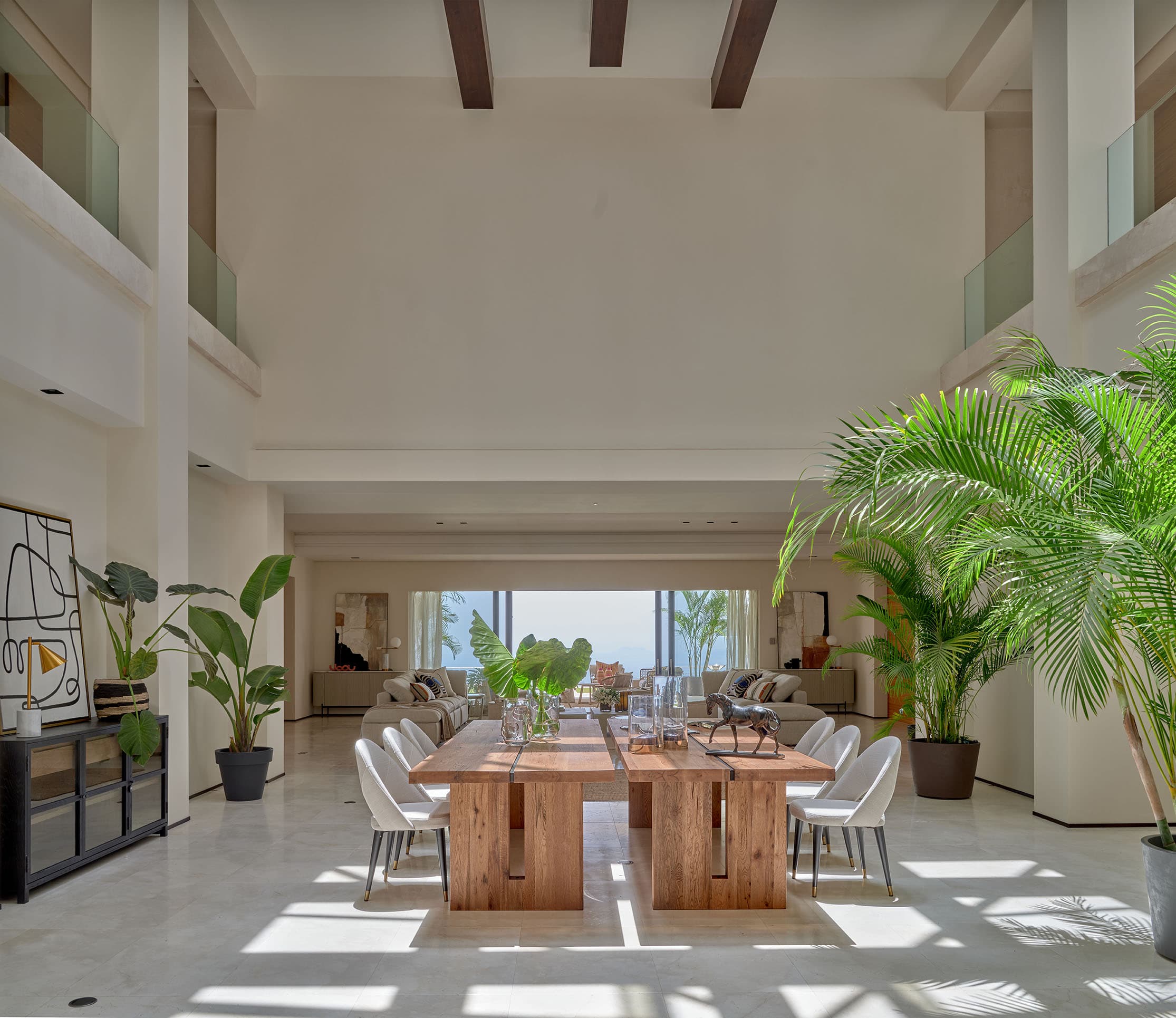 Vista general del gran comedor con galería en planta superior de vidrio, mesa de madera de tablones con sillas blancas y abundante vegetación tropical en el interior de la villa.