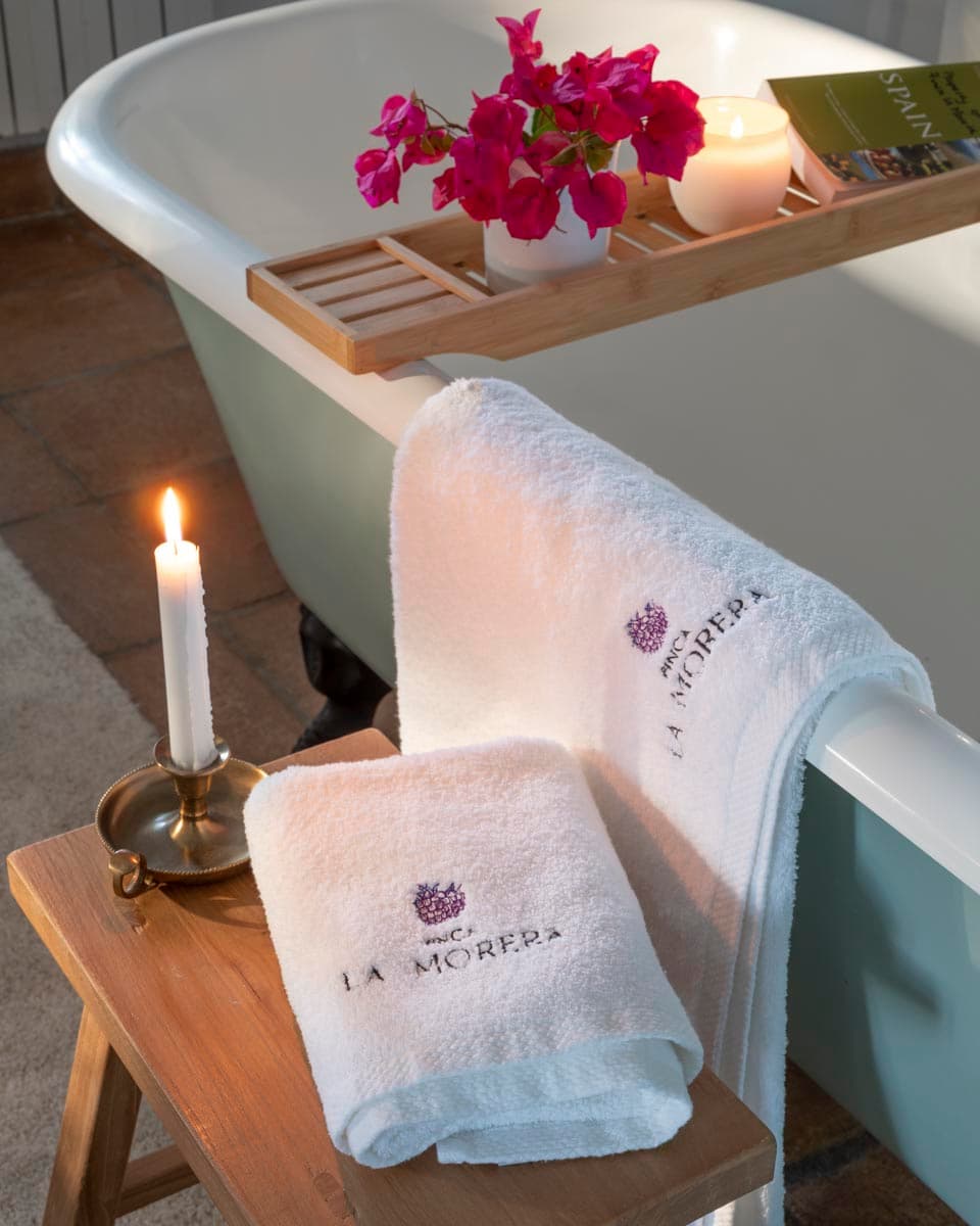 Primer plano de toallas blancas con el logotipo bordado de Finca La Morera, apoyadas en un taburete de madera junto a la bañera exenta azul. Buganvilla rosa y una vela encendida completan la escena de lujo boutique.