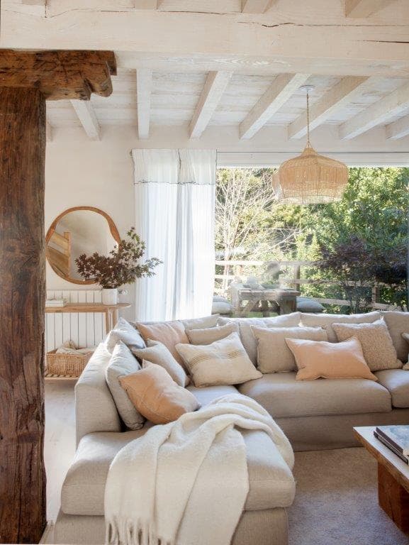 Rincón del salón con sofá rinconera en lino beige claro, lámpara de rafia y viga de madera rústica oscura en primer plano. Al fondo se aprecia una consola con espejo de forma orgánica y decoración de ramas secas.