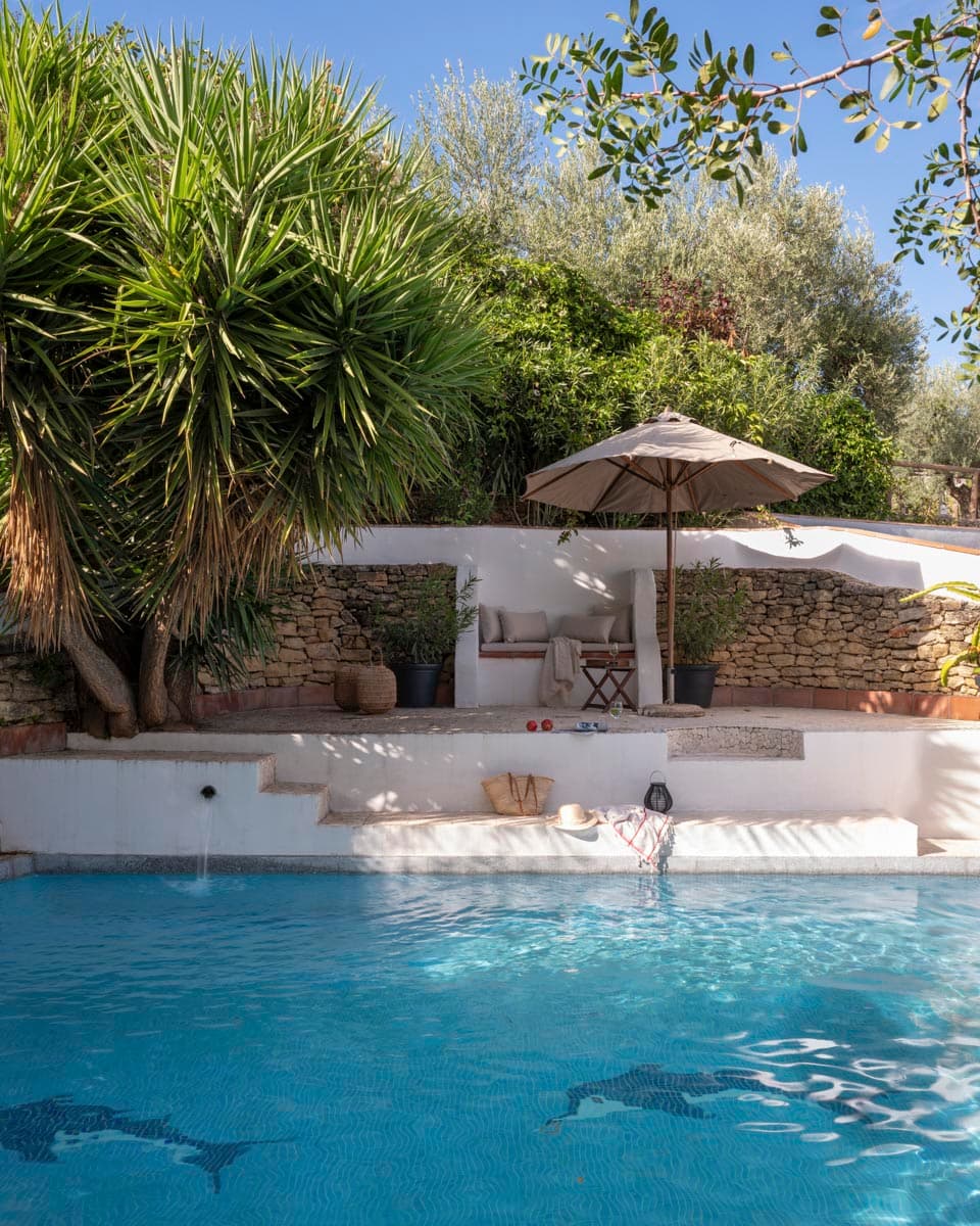 Piscina con agua turquesa y figura de delfín en mosaico en el fondo, rodeada de piedra natural. Un banco con sombrilla, palmeras y olivos enmarcan la piscina en un paisaje mediterráneo de verano.