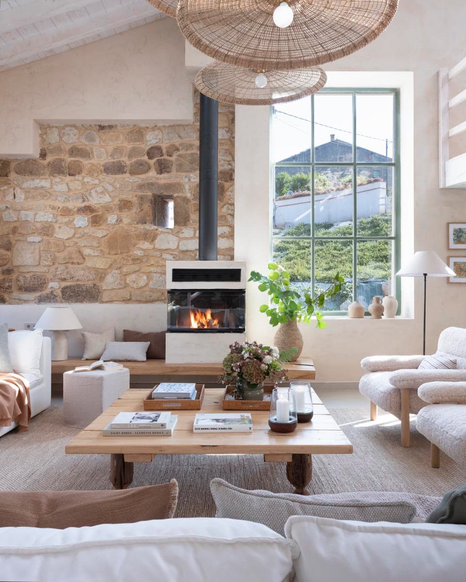 Vista cercana del salón con imponente muro de piedra natural, chimenea de leña encendida y gran ventanal verde que enmarca el paisaje exterior, con mesa de café rústica y butacas de tejido bouclé.