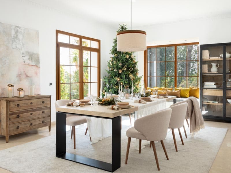 Comedor con mesa rectangular de madera sobre base metálica negra y sillas tapizadas beige, con el árbol de Navidad decorado como telón de fondo y el cuadro abstracto rosado a la izquierda. La lámpara de tambor de doble tono y la vitrina negra lateral completan un comedor de línea contemporánea.