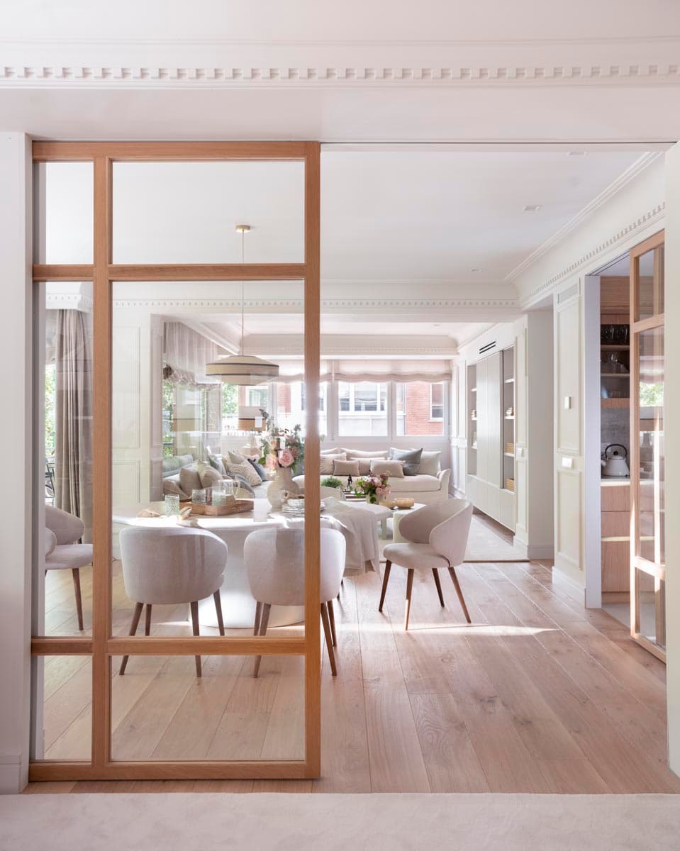 Mampara corredera de roble natural y cristal en primer plano que divide el pasillo del comedor; a través del cristal se ve la mesa ovalada blanca con flores rosas y, al fondo, los sofás del salón bañados de luz natural. La continuidad de las molduras clásicas en techo unifica los espacios.