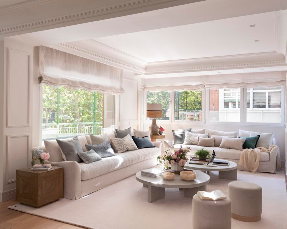 Vista frontal del salón con dos sofás de funda de lino blanca con abundantes cojines en gris azulado y verde oscuro, mesas de centro redondas y puf de terciopelo gris. Los estores de lino con ondas enmarcan los amplios ventanales con vistas a los árboles de la calle, mientras una mesa auxiliar de madera maciza añade calidez.