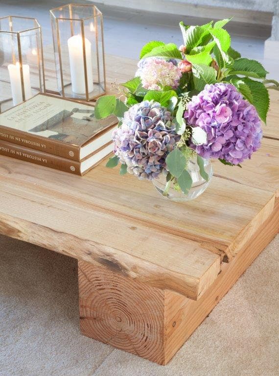 Primer plano de la mesa de centro de madera maciza con un ramo de hortensias moradas y azules en jarrón de cristal, faroles metálicos con velas y libros apilados. El acabado artesanal de la madera con su veta natural es el protagonista de la imagen.