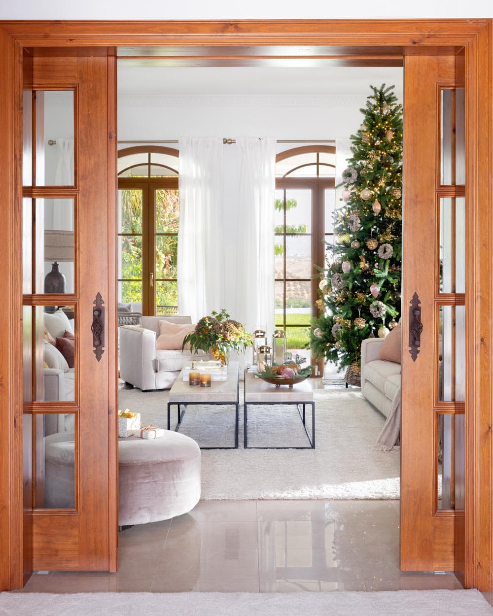 Vista desde el vestíbulo a través de puertas correderas de madera con vidrio hacia el salón luminoso, donde el árbol de Navidad destacado con luces cálidas y adornos rosas y dorados preside el espacio. El contraste entre la madera cálida de las puertas y los sofás blancos crea una imagen de sofisticada elegancia festiva.