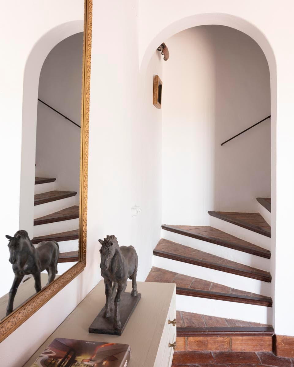 Escalera interior con peldaños de barro y paredes blancas encuadrada por un arco de medio punto. En primer plano, aparador con escultura de caballo en bronce reflejada en el gran espejo dorado. Arquitectura andaluza tradicional.