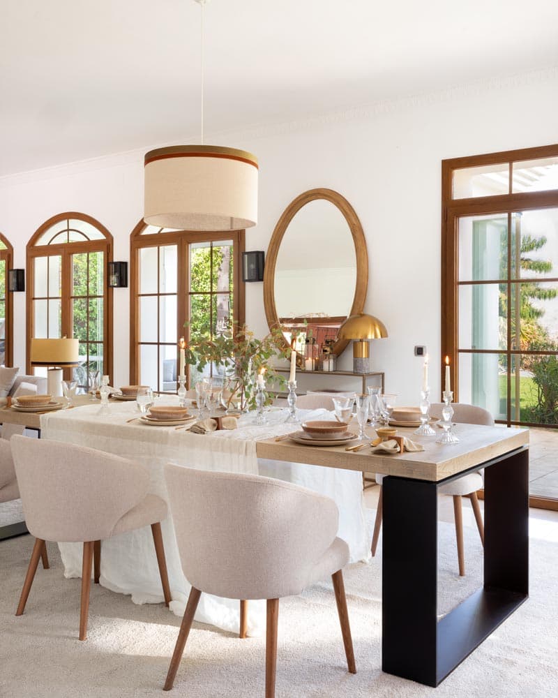 Mesa rectangular con centro de ramas naturales, candelabros de cristal y vajilla nude-beige, con el espejo oval dorado al fondo sobre la consola y la lámpara de tambor colgante. Las ventanas arqueadas con la luz vespertina crean una atmósfera cálida e íntima para una cena elegante.