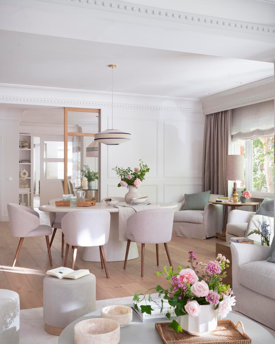 Vista desde el salón hacia el comedor: en primer plano la mesa de centro redonda con ramo de flores rosas, al fondo la mesa ovalada con sillas tapizadas en blanco roto y la lámpara colgante de lino con franja azul colgada sobre latón dorado. La mampara de roble al fondo conecta visualmente con la cocina y el salón de lectura.