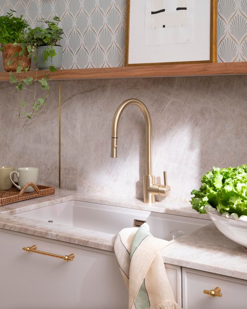 Primer plano del fregadero de cocina empotrado en encimera de mármol gris veteado, con grifo de cuello de cisne en latón dorado mate y tiradores en latón en los cajones. En el estante de madera superior descansan plantas colgantes y un cuadro enmarcado en dorado, frente al papel pintado con motivo de abanico art déco.