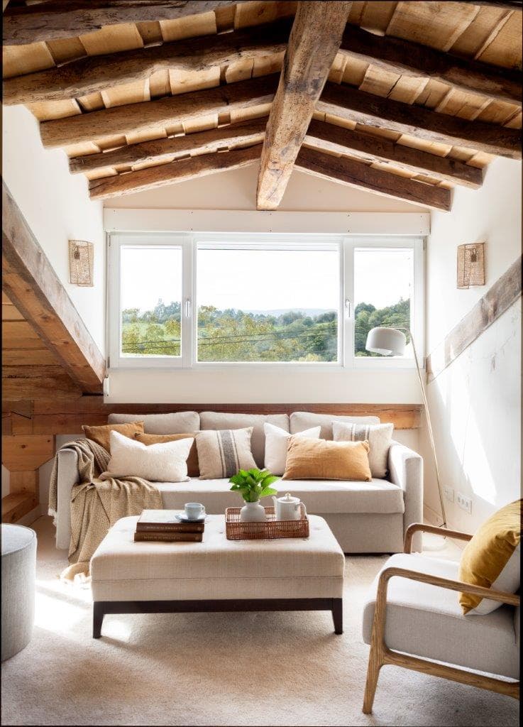 Acogedora sala de estar en la buhardilla con techos inclinados de madera rústica, sofá y sillón tapizados en lino, puf cuadrado de tela y vistas al campo a través del ventanal. Las apliques de rafia y el sillón de madera con cojín mostaza completan el espacio.