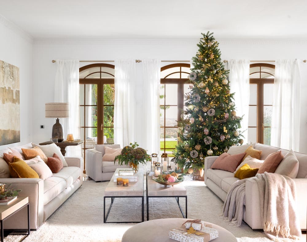 Amplio salón luminoso con sofás en tonos crema y cojines en rosa y mostaza, presidido por un majestuoso árbol de Navidad decorado en dorado y rosa junto a ventanas con arcos de madera. La decoración navideña se integra con elegancia en un ambiente cálido y sofisticado.
