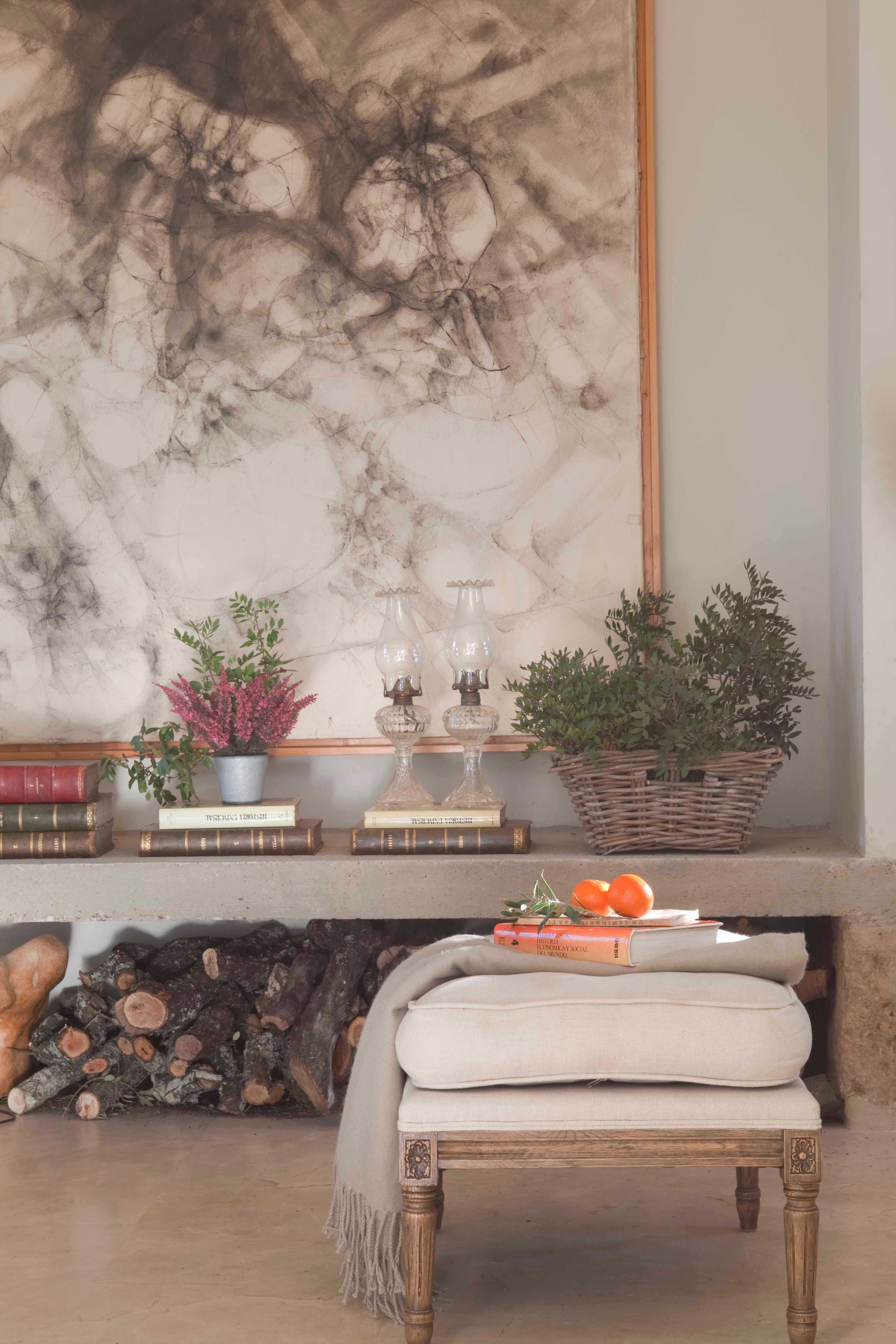 Detalle de un rincón donde una gran obra de arte abstracta en carboncillo preside el espacio sobre un estante de cemento con libros antiguos, lámparas de petróleo de cristal y plantas. Bajo el estante se apila leña. En primer plano, un taburete de estilo Luis XVI tapizado en lino con libros y naranjas.
