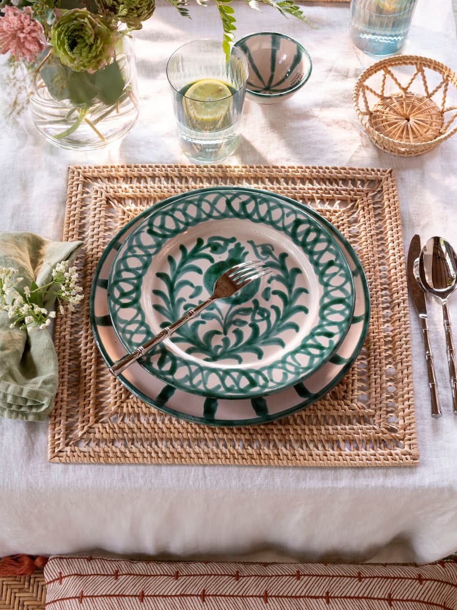 Primer plano de un cubierto individual sobre bajoplato cuadrado de fibra trenzada. La vajilla artesanal en blanco con cenefa en verde botella —plato llano con motivos vegetales y cuenco a rayas— se combina con servilleta de lino verde salvia doblada, cubiertos plateados y vasos de vidrio soplado. La estética es artesanal, natural y con marcado carácter mediterráneo.