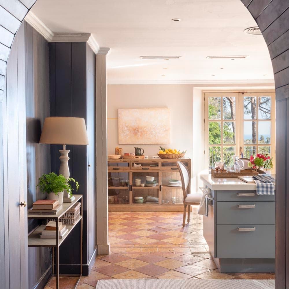 Vista desde el corredor a través de un arco pintado en gris antracita hacia la cocina-office, con una consola con lámpara y plantas a la izquierda y la isla de cocina en gris-verde y ventana al jardín a la derecha. El suelo de terracota en diamante unifica ambos espacios.