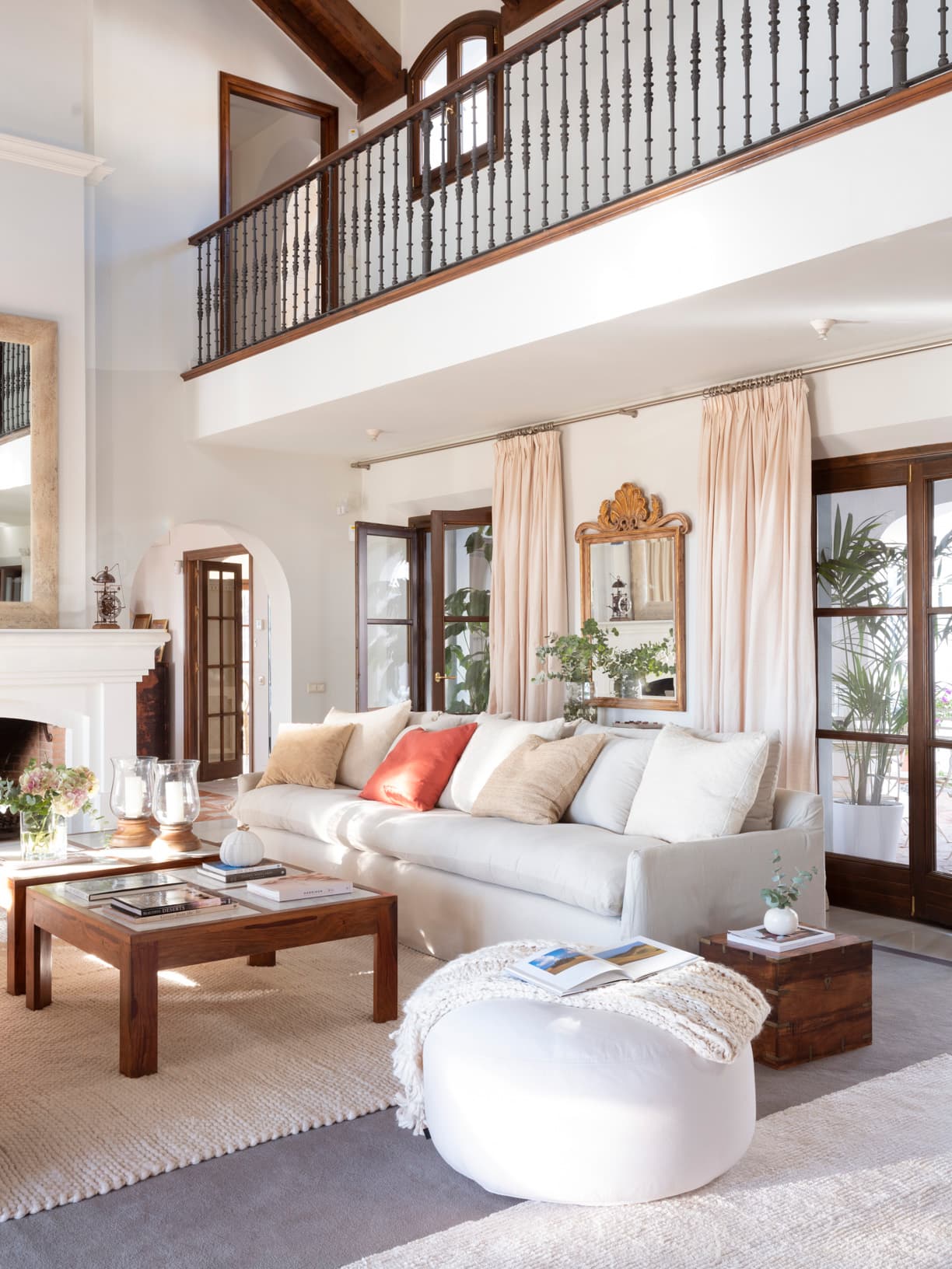 Salón de doble altura con sofá de lino blanco, mesa ratona de madera oscura y puf de punto. La chimenea blanca con espejo de marco dorado tallado en estilo barroco es el centro visual. Una galería con balaustre de hierro y ventanas con palmeras al fondo definen el carácter mediterráneo de la estancia.