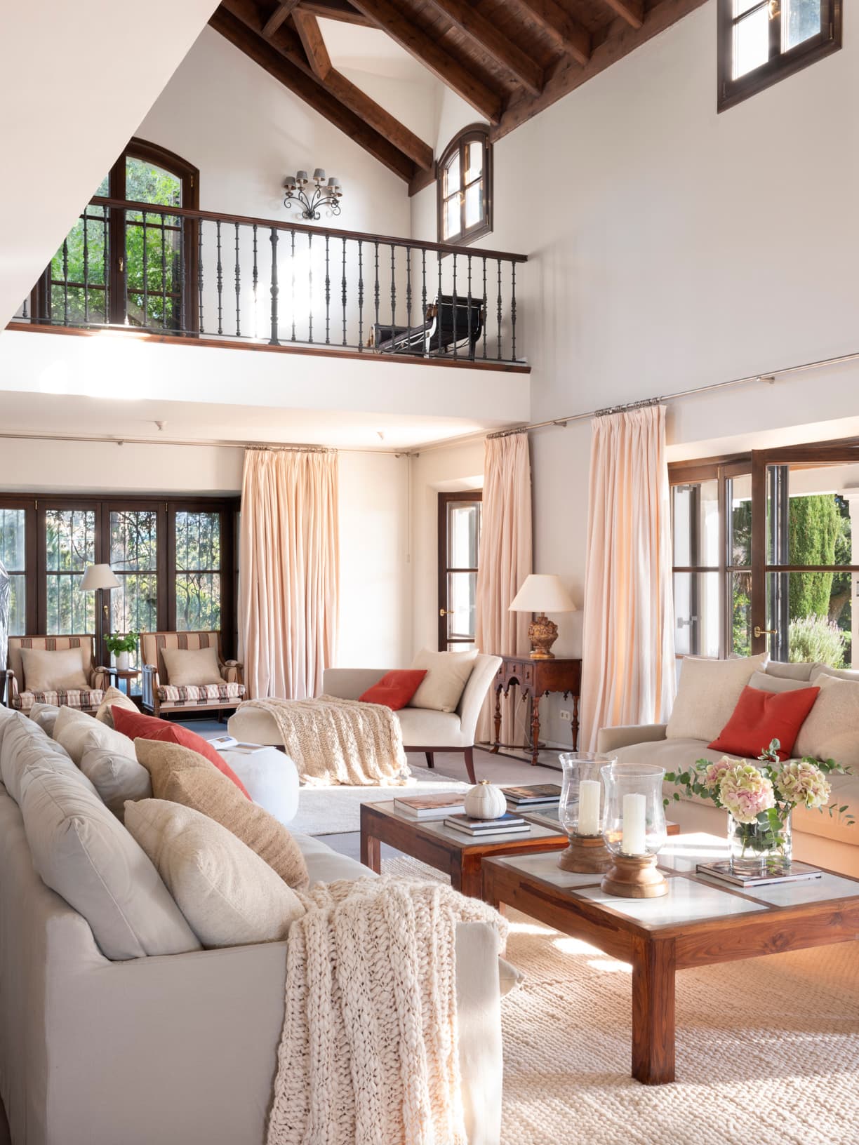 Salón de doble altura con vigas de madera oscura y galería con balaustre de hierro forjado. Sofás beige y crema con almohadones, espejo dorado de estilo barroco, mesas de madera oscura y grandes ventanas y puertas con vistas al jardín. La luz natural inunda el espacio con un ambiente de elegancia campestre.