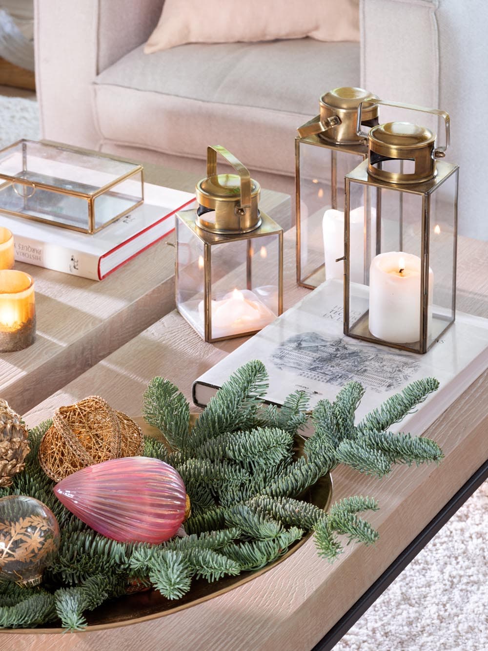 Detalle de la mesa de centro con bandeja dorada que alberga ramas de abeto azul, bolas navideñas rosas y doradas, junto a farolillos de cristal y latón dorado con velas encendidas y libros de decoración apilados. La composición combina lo festivo con lo cotidiano de forma equilibrada.