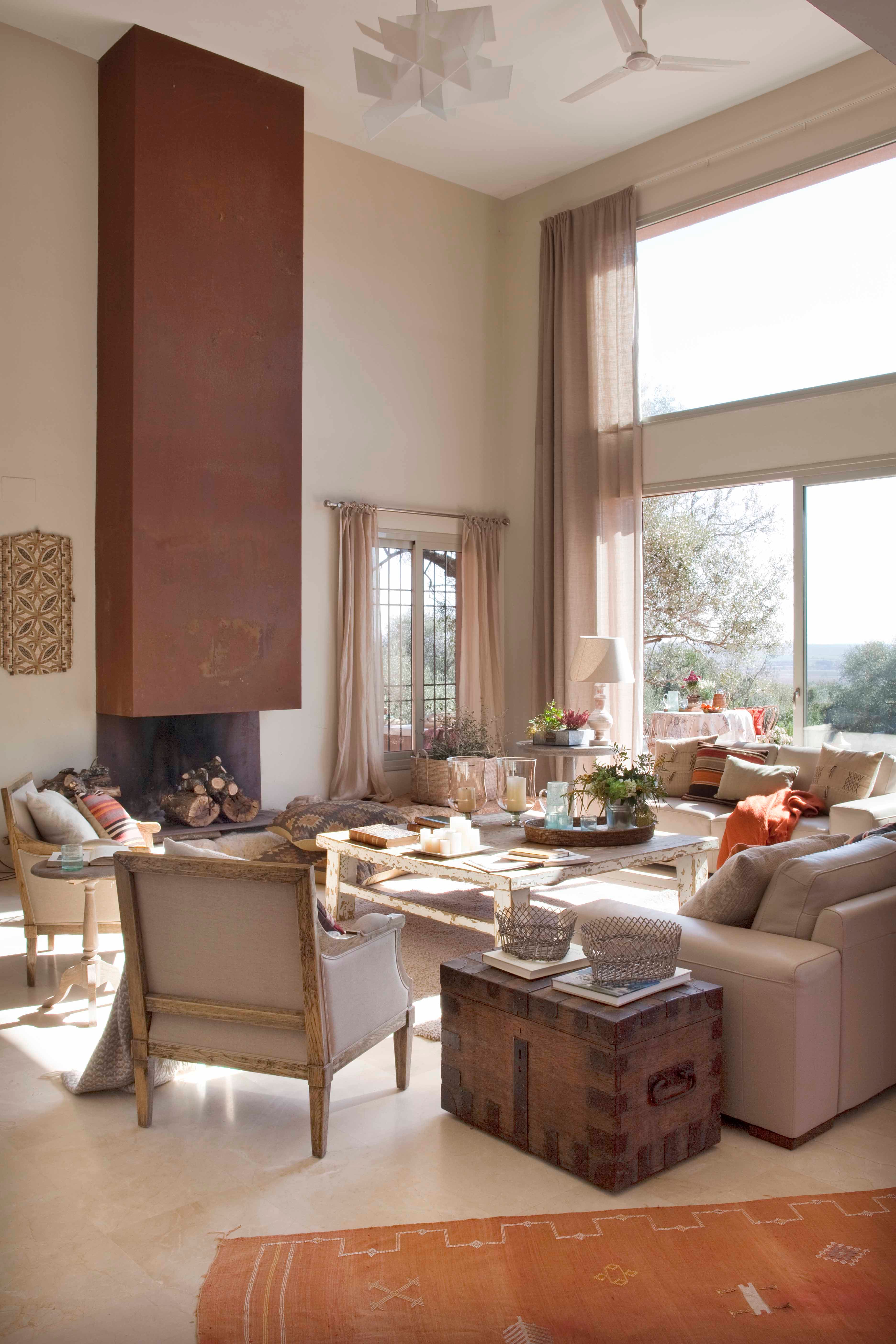 Salón de doble altura con chimenea de acero corten y gran ventanal panorámico con vistas al jardín y paisaje. La decoración combina sofás de lino, sillones de madera envejecida tapizados en lino, una mesa ratona rústica, baúl vintage y alfombra kilim naranja. Ventilador de techo y aplique decorativo de madera tallada completan el espacio.