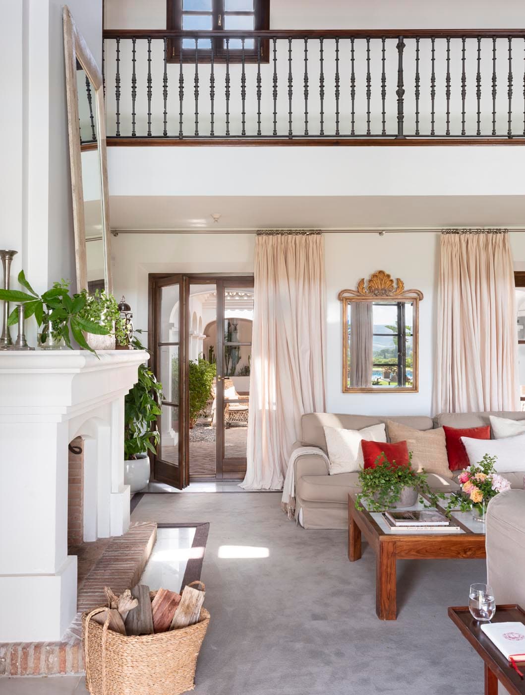 Salón claro con sofás blancos de lino, chimenea de mármol blanco con espejo dorado de estilo barroco sobre la repisa y cortinas en rosa pálido. El balcón de hierro forjado en el piso superior y las puertas francesas al fondo enmarcan el espacio con nobleza arquitectónica.