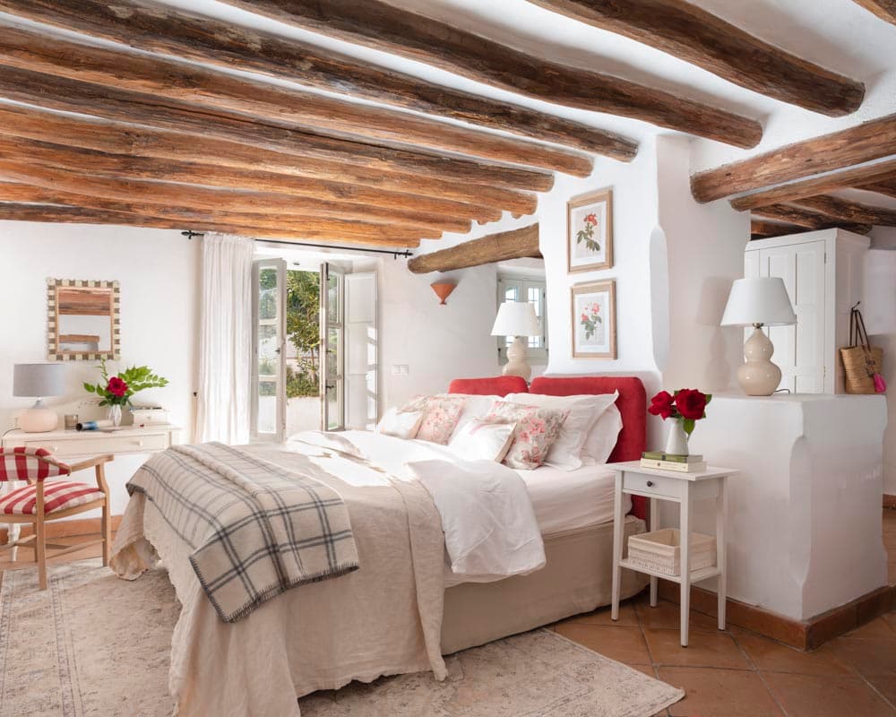 Dormitorio principal con techumbre de vigas de madera oscura expuesta, cabecero tapizado en rojo y ropa de cama floral. Un escritorio blanco, silla a cuadros, mesita de noche y ampias puertas al jardín completan la estancia de carácter rústico.