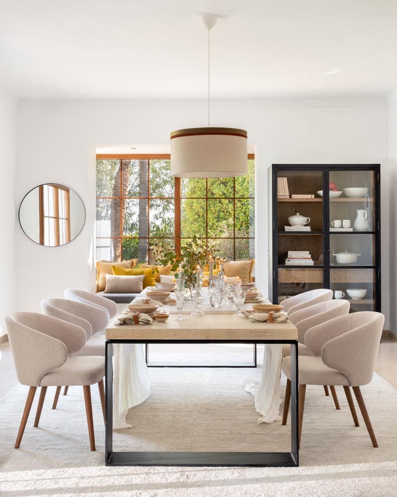 Comedor con mesa rectangular de madera clara sobre estructura negra y sillas tapizadas beige, con un banco-ventana con cojines mostaza al fondo y una elegante vitrina negra con menaje blanco. La lámpara de tambor bicolor y el espejo circular pequeño completan un comedor contemporáneo de gran coherencia estética.
