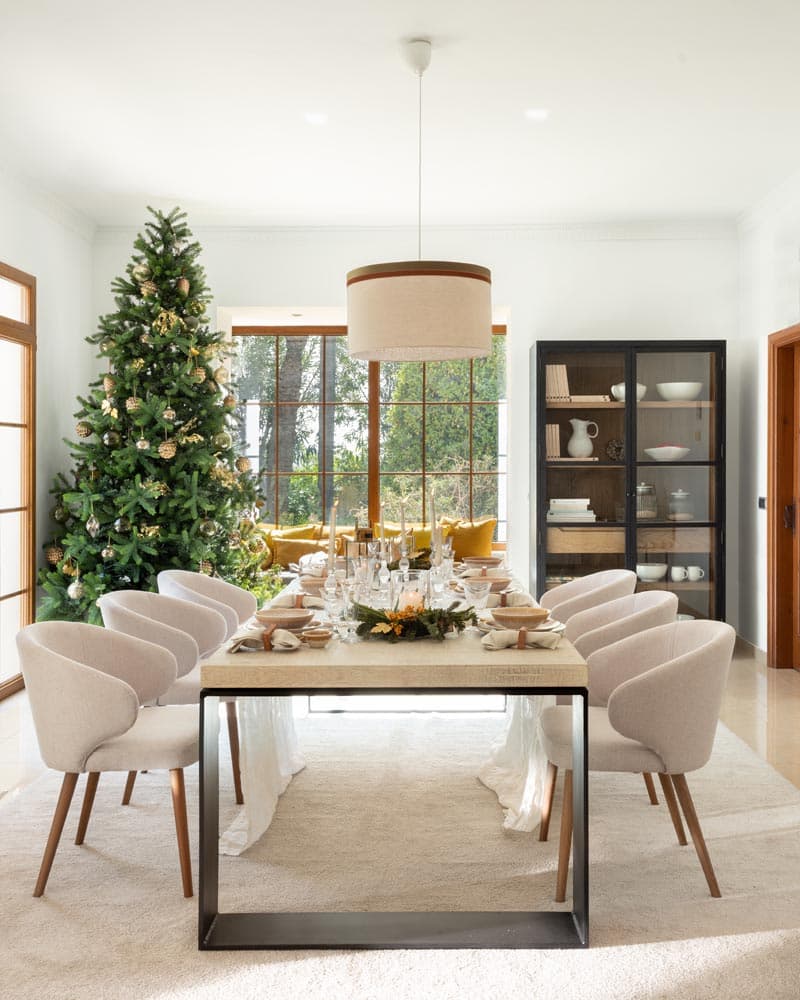 Vista frontal de la mesa rectangular vestida para Navidad, con mantel blanco, vajilla elegante, centro de mesa floral y un gran árbol iluminado junto a la ventana con vistas al jardín.