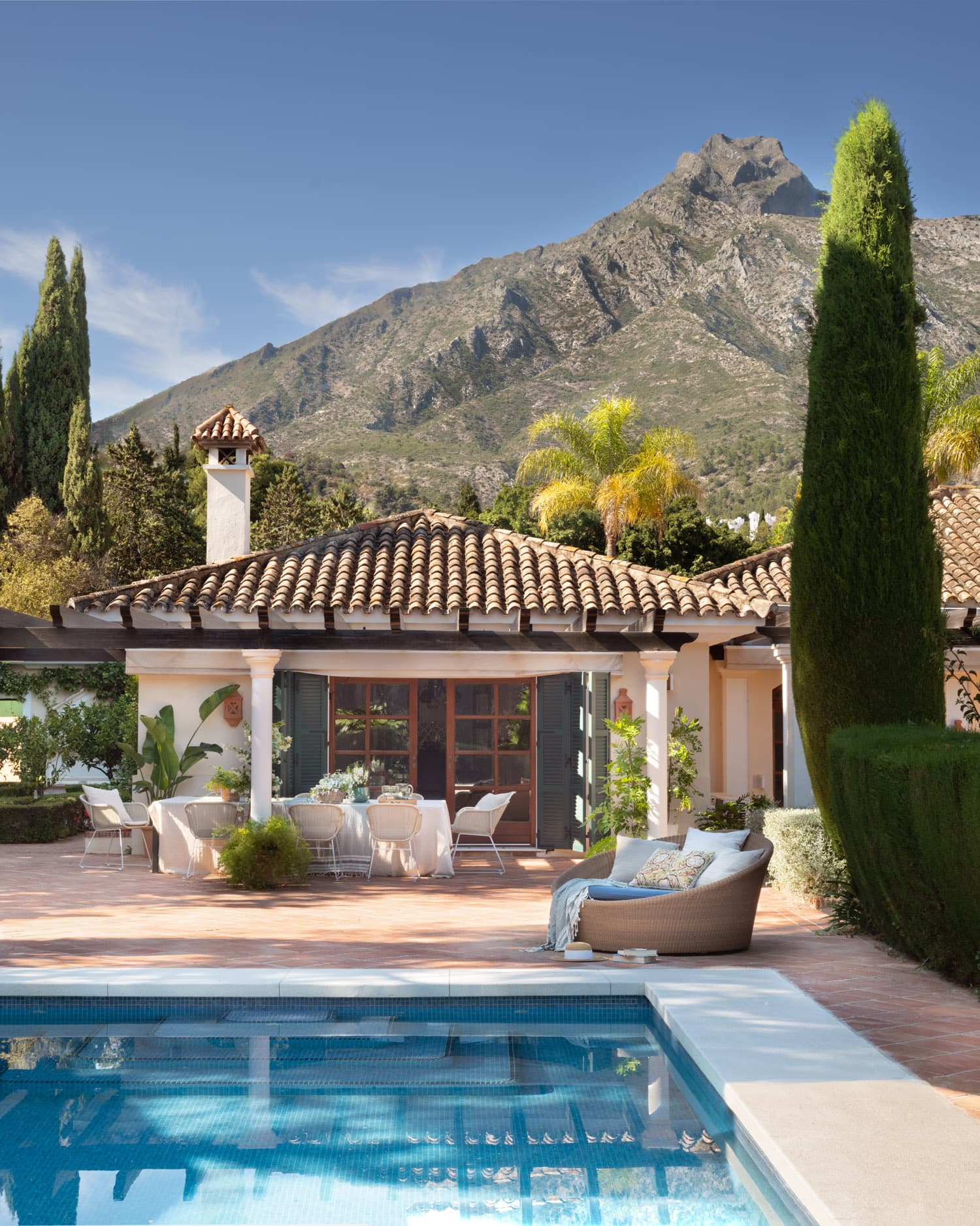 Vista de la piscina exterior con agua azul cristalina en primer plano, el porche con mobiliario de exterior blanco y el tejado mediterráneo de teja árabe de la vivienda al fondo. Los cipreses italianos y las palmeras flanquean la fachada, y la imponente silueta de la montaña se alza bajo un cielo de verano perfectamente azul.