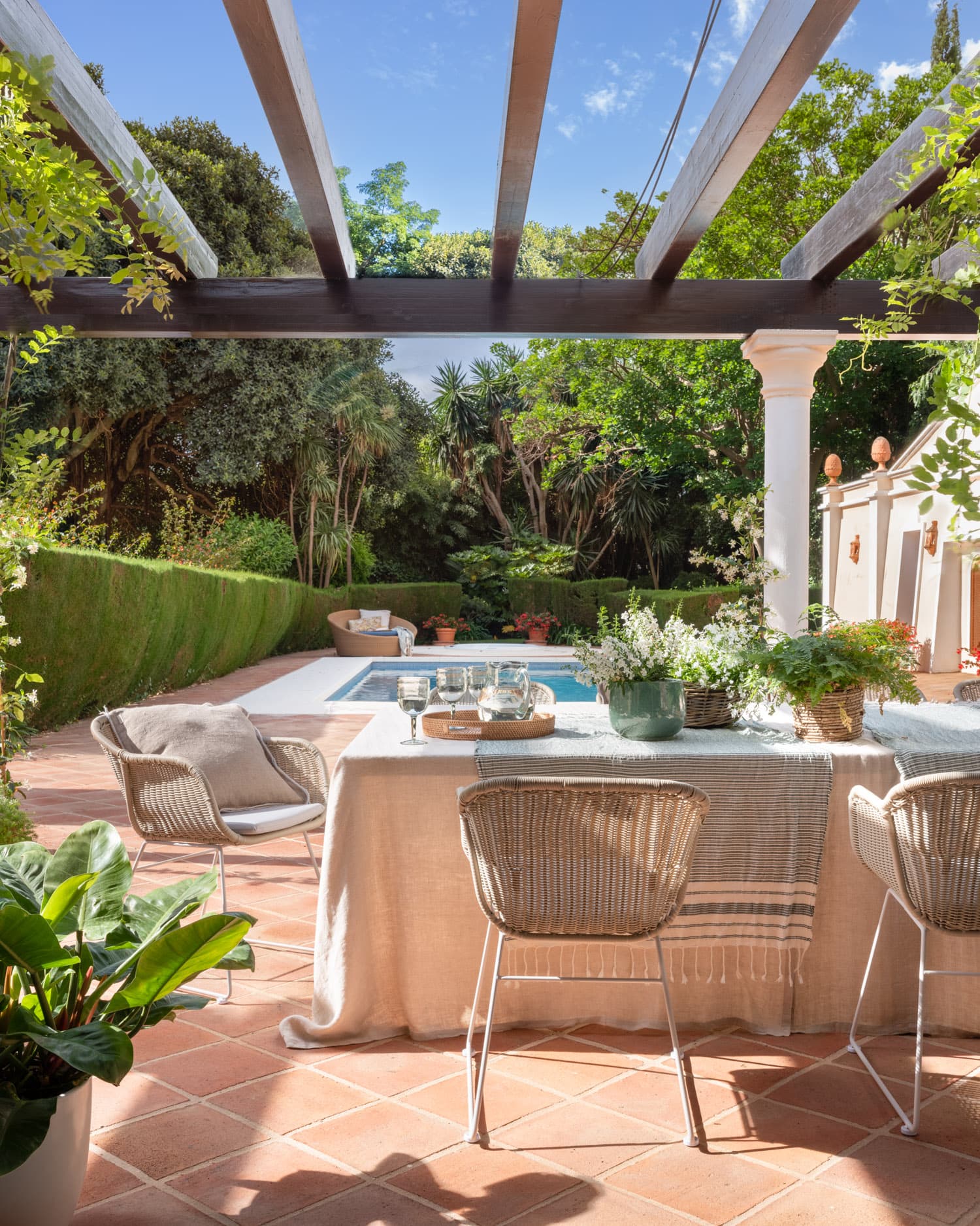 Rincón de comedor exterior íntimo bajo una pérgola de vigas de madera oscura. Una mesa vestida con mantel de lino y sillas de ratán de fibra natural se sitúa junto a la piscina, con el jardín tropical al fondo. La decoración floral con plantas aromáticas y cristalería delicada aporta un ambiente fresco y sofisticado.