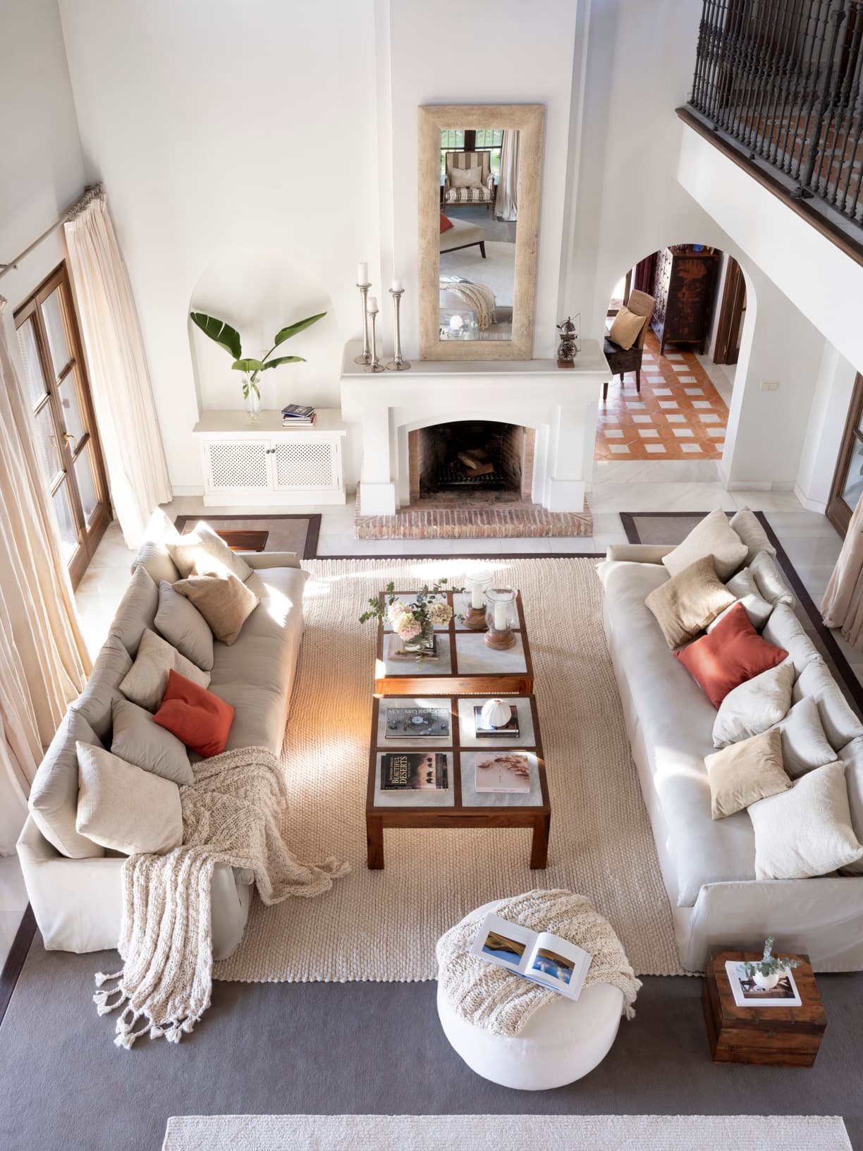 Vista cenital del salón que muestra la distribución simétrica de los sofás de lino alrededor de la chimenea blanca. Una mesa ratona de madera con cristal en el centro y puf de punto blanco. Al fondo, el acceso a otras estancias con arco en ladrillo visto. La galería superior con balaustre enmarca la composición.