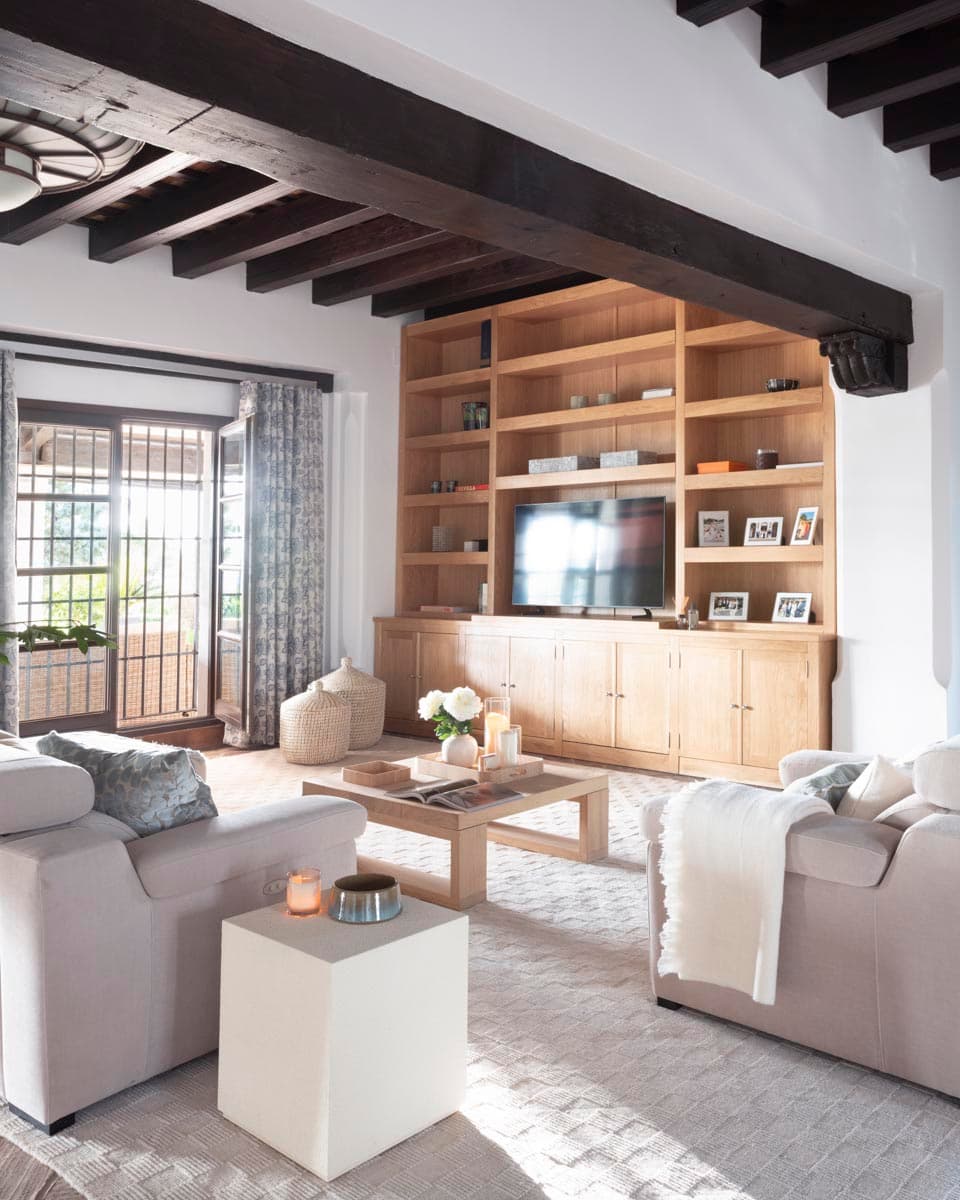 Zona de estar con gran librería empotrada de madera de roble natural que aloja la televisión, flanqueada por sofás grises y mesa de café de madera. Vigas oscuras al techo y ventana con reja y cortinas estampadas con luz natural.