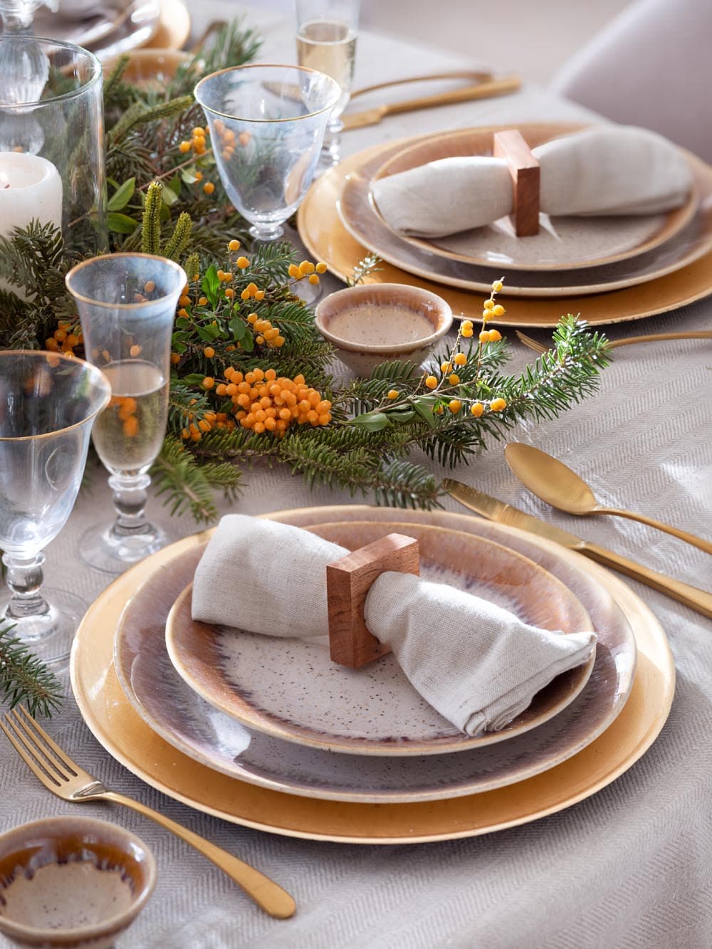 Detalle de la mesa navideña con vajilla artesanal en cerámica degradada rosa-gris sobre plato bajo dorado, cubiertos dorados y copas de cristal soplado, junto a un centro con ramas de pino, bayas naranjas y velas. La servilleta de lino se anuda con un soporte en madera natural.