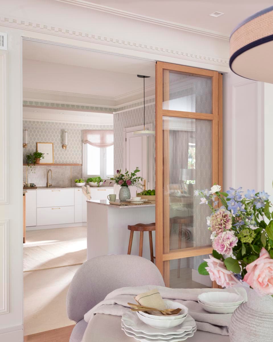Desde el comedor, con silla tapizada en primer plano, se contempla la cocina blanca con isla de encimera de mármol y taburetes de madera natural, separada por una vidriera de roble con cristal translúcido. El papel pintado geométrico en gris pálido y las apliques dorados dan carácter al espacio.