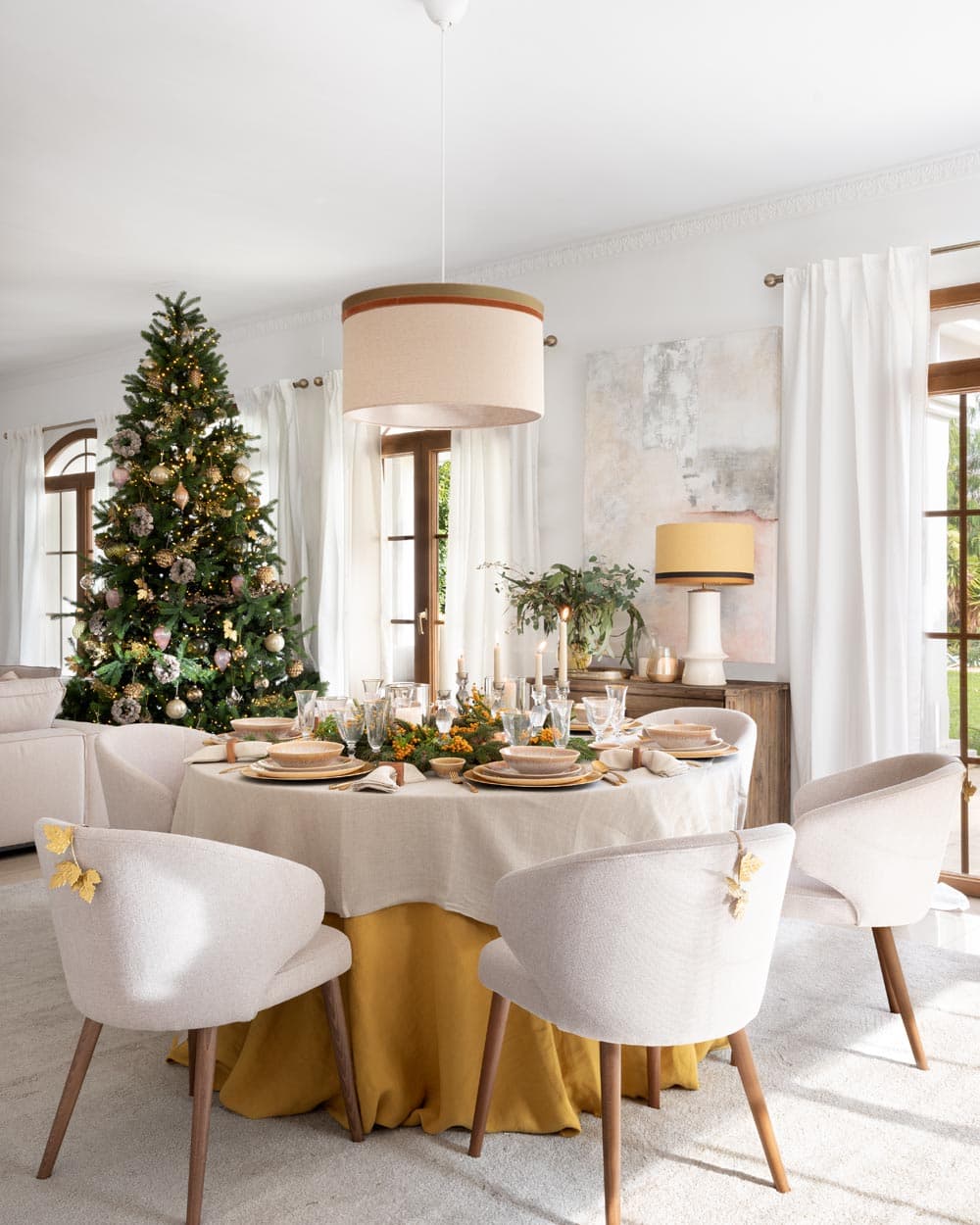 Vista general del espacio de comedor abierto al salón, con el gran árbol de Navidad decorado en rosa y dorado al fondo junto a lienzo abstracto en tonos neutros. La mesa redonda vestida combina con las sillas de estilo contemporáneo y la cómoda de madera rústica con lámpara de cerámica.