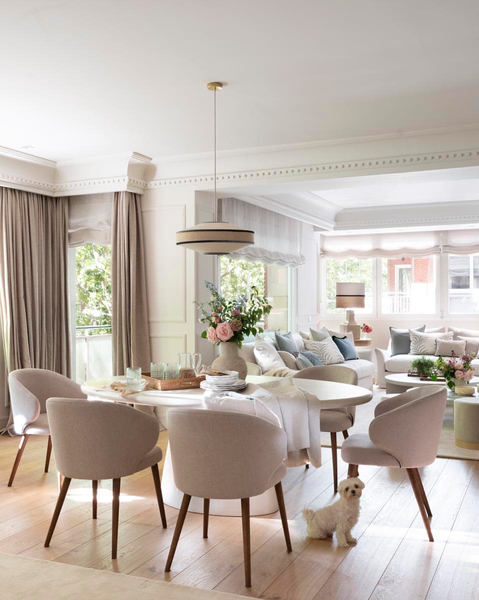 Amplio salón comedor con mesa ovalada blanca rodeada de sillas tapizadas en beis y patas de madera, lámpara colgante de lino con detalle en azul marino y suelo de roble natural. Al fondo, el salón se abre con sofás blancos y cojines en tonos azul y gris, con molduras clásicas en techo y paredes.