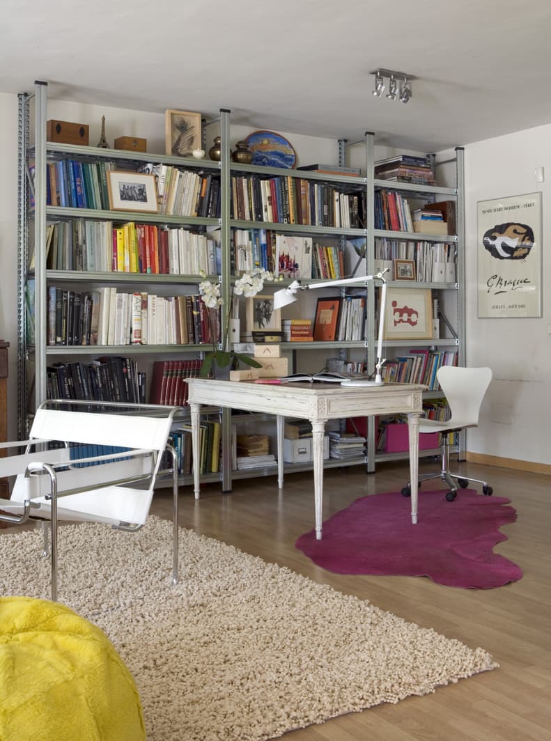 Vista general del despacho-biblioteca con estanterías metálicas industriales que cubren toda la pared del fondo, cargadas de libros y objetos decorativos. La mesa de escritorio provenzal lacada en blanco ocupa el centro, con una silla de oficina blanca. Una silla Wassily en blanco y cromado se sitúa a la izquierda sobre la alfombra de pelo largo en beige. En primer plano inferior izquierdo aparece un puf amarillo vibrante que aporta contraste. La alfombra de cuero fucsia delimita el área de trabajo. El suelo es de parquet de roble claro.