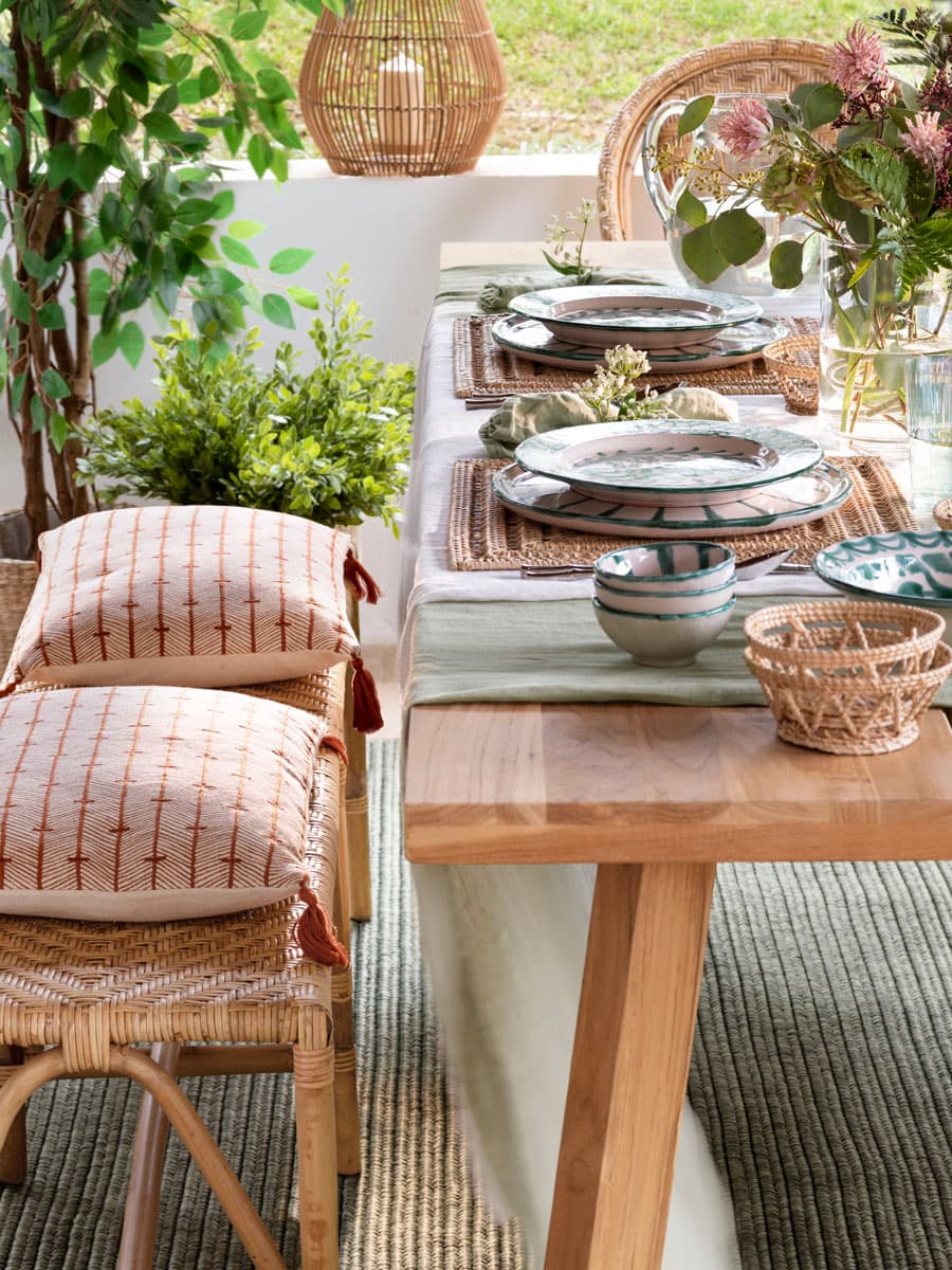 Vista lateral de la mesa de comedor exterior bajo el porche, con tablero de madera maciza natural y patas en caballete. Las sillas de ratán natural acolchadas con cojines de tela estampada en terracota y arena enmarcan la mesa. En primer plano, un taburete de ratán con almohadones de lino estampado en rojo ladrillo sobre fondo crudo. Al fondo, la apertura del porche al jardín con palmeras y vegetación tropical.