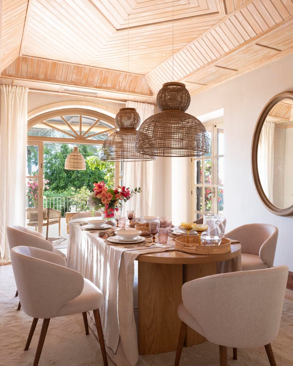 Comedor luminoso con mesa oval de madera natural vestida con mantel de lino y vajilla rosa-polvorienta, dominado por tres grandes lámparas de ratán escalonadas bajo un espectacular techo octogonal de madera clara. Las ventanas arqueadas abiertas al jardín inundan la estancia de luz natural.