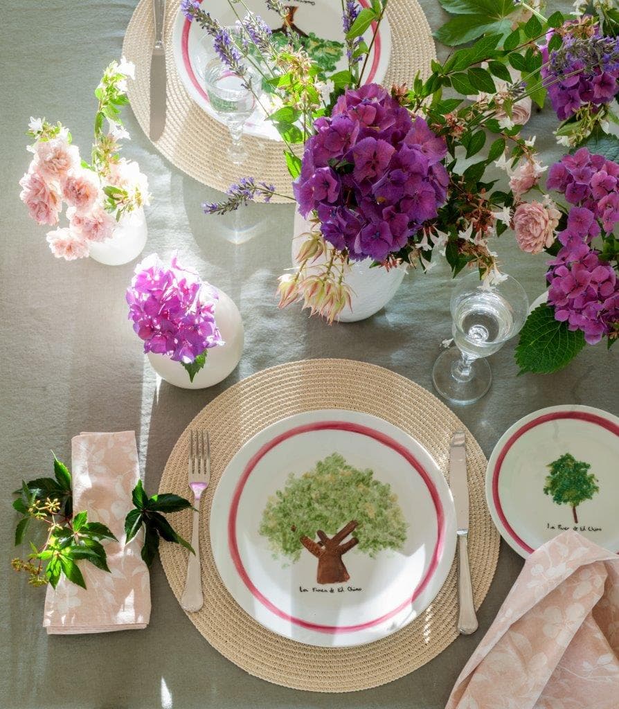 Detalle muy cuidado de un puesto individual con plato artesanal de cerámica con ilustración de árbol en verde acuarela con borde rojo —firmado 'La Finca de El Olmo'— sobre salvamanteles de esparto. Junto a él, jarrones blancos con hortensias moradas, rosas de jardín y flores silvestres.