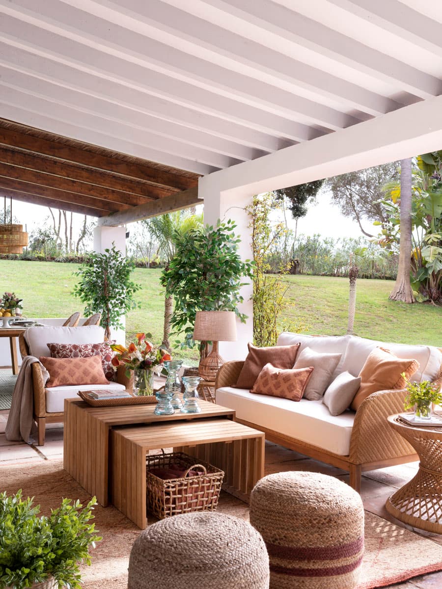 Vista de la zona de estar del porche cubierto con techo de madera pintado en blanco y vigas de madera natural vistas. El conjunto de sofás de ratán con tapizado en crudo y cojines en tonos terracota, salmón y tierra se organiza en torno a una mesa de centro de madera maciza en formato nesting. Puffs de esparto rayados y una mesa lateral de ratán completan el mobiliario. Plantas de interior de gran porte —ficus, monstera— aportan frescura y conexión con el entorno natural.