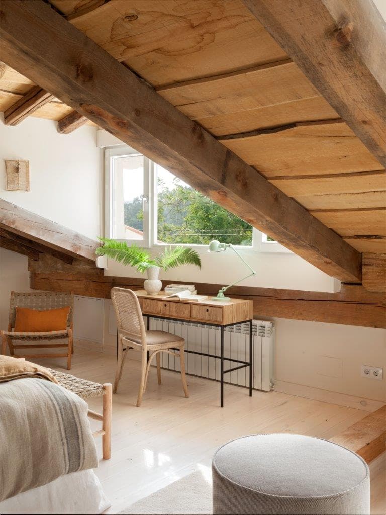 Dormitorio en buhardilla con vigas de madera rústica al descubierto y techos inclinados, escritorio de madera con patas metálicas y silla de rejilla, y helechos como decoración verde. La lámpara articulada en verde menta y la silla de ratán aportan detalles vintage.